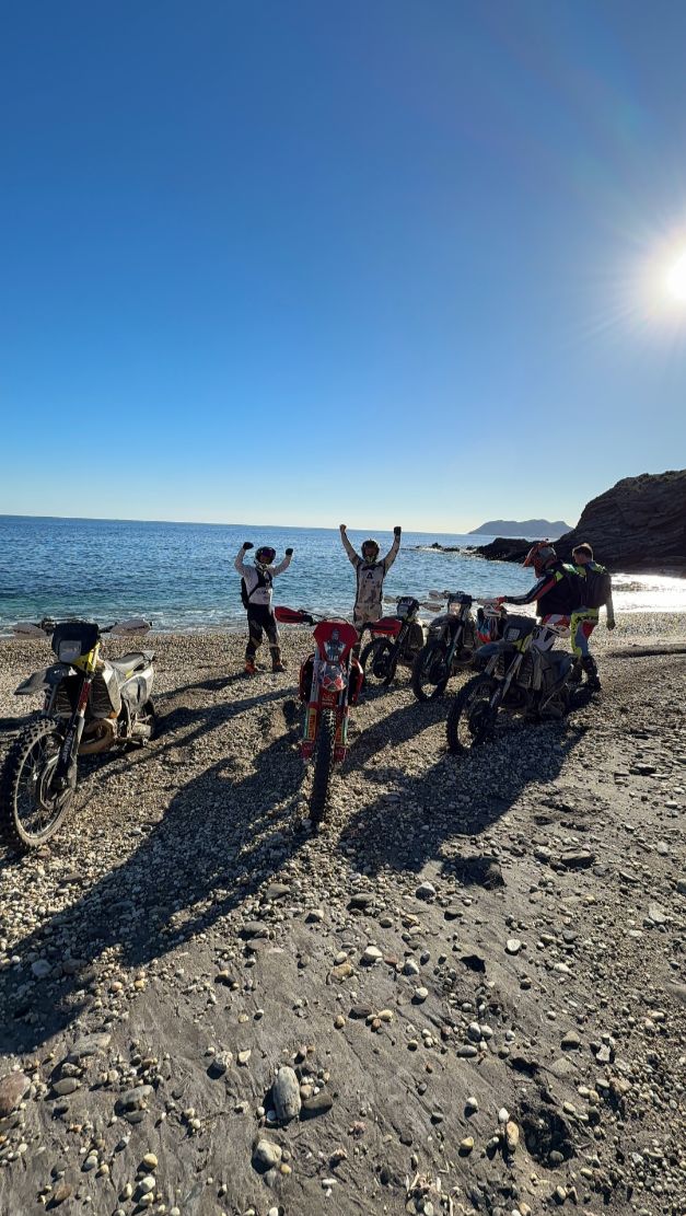 Enduro riders celebrating with their bikes on a pebble beach on the Mediterranean
