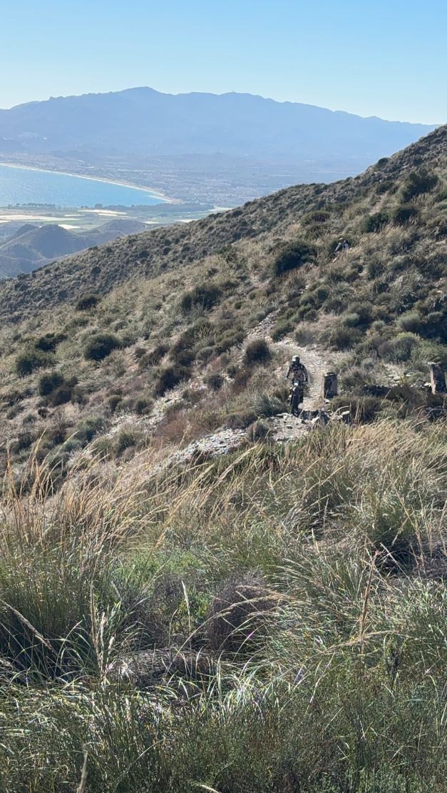Riders on a mountainside singletrack with the sea and coastal mountains behind