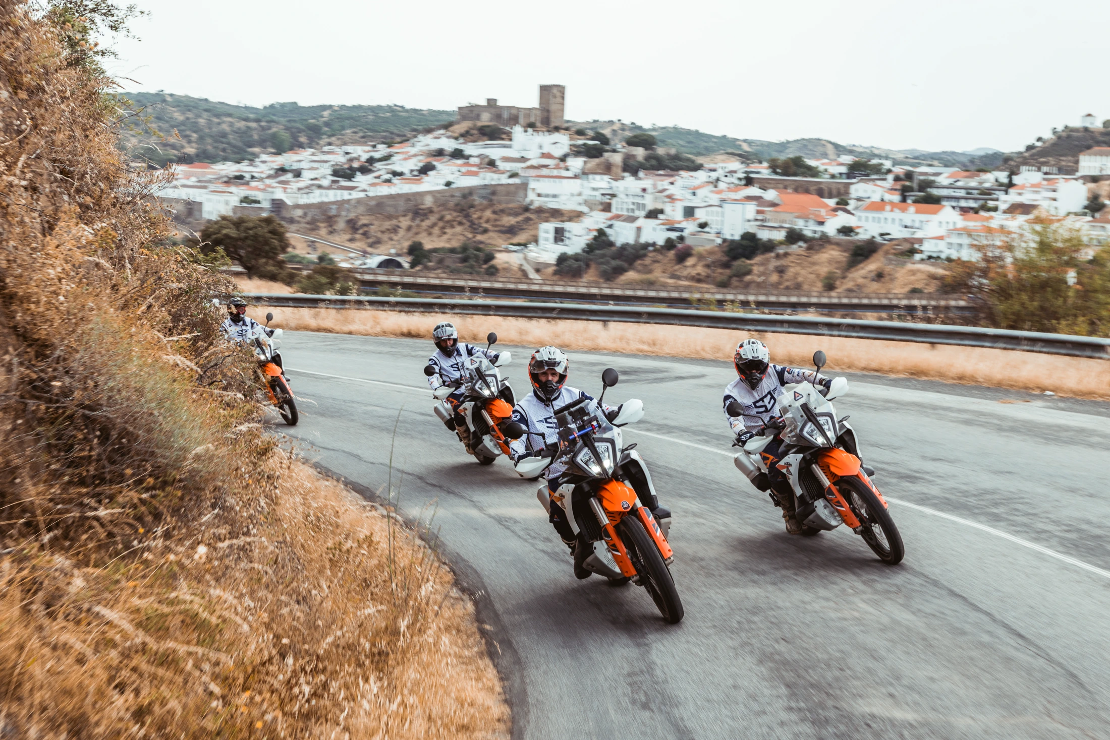 Riders on KTM adventure bikes on a road bend below a white village with a castle