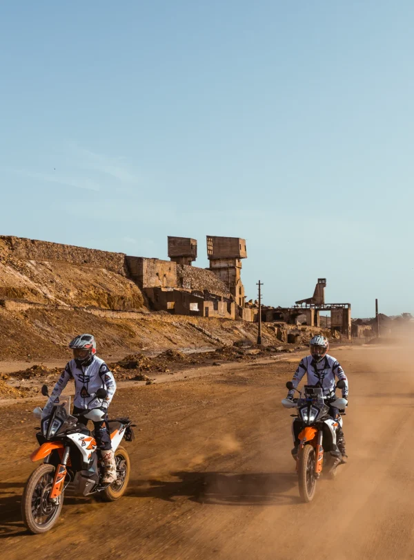 Riders on KTM adventure bikes on a dusty track past abandoned mine buildings