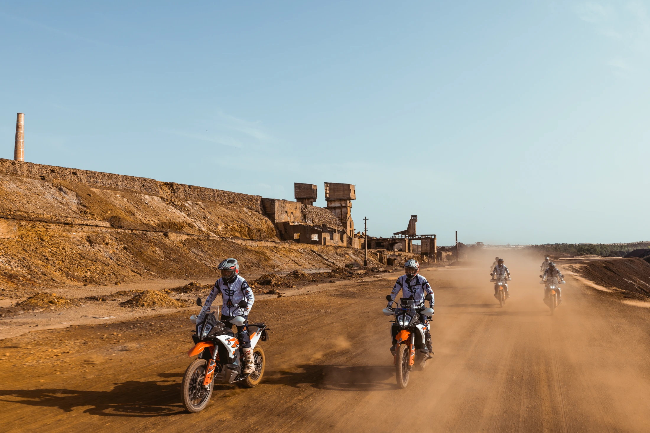 Riders on KTM adventure bikes on a dusty track past abandoned mine buildings