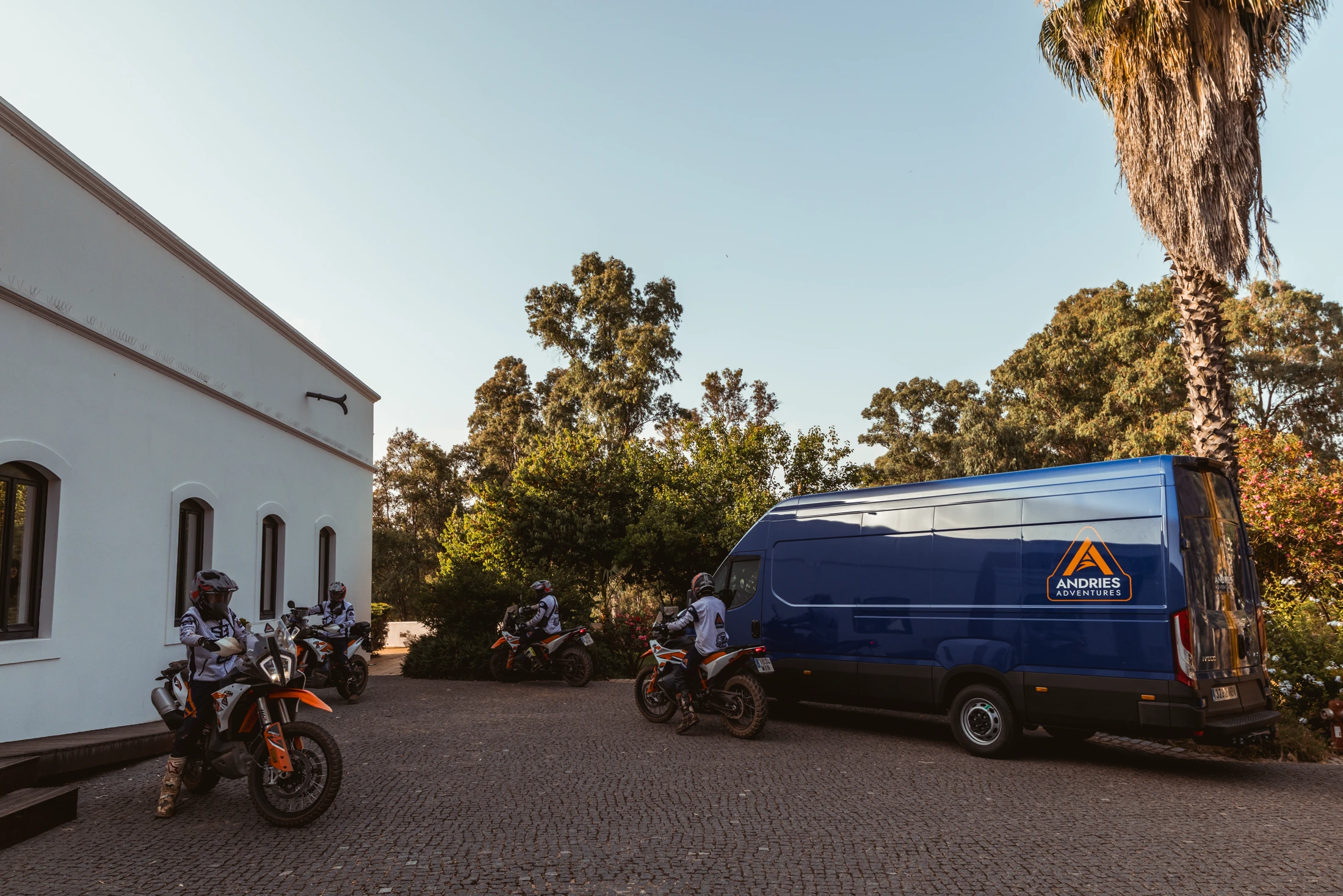 Riders and KTM adventure bikes next to the Andries Adventures support van at a white hotel