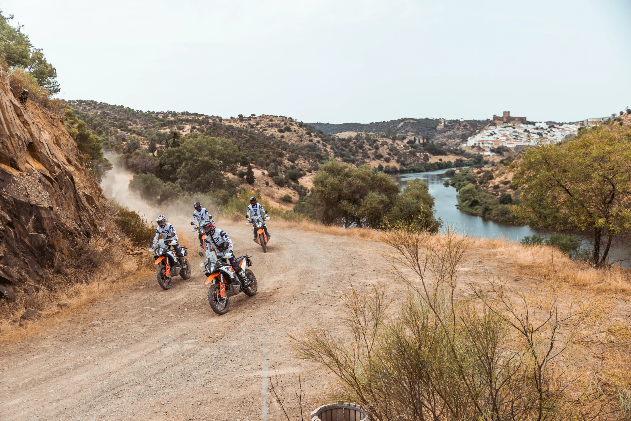 Riders on KTM adventure bikes on a dirt track above a river valley with a white hilltop town