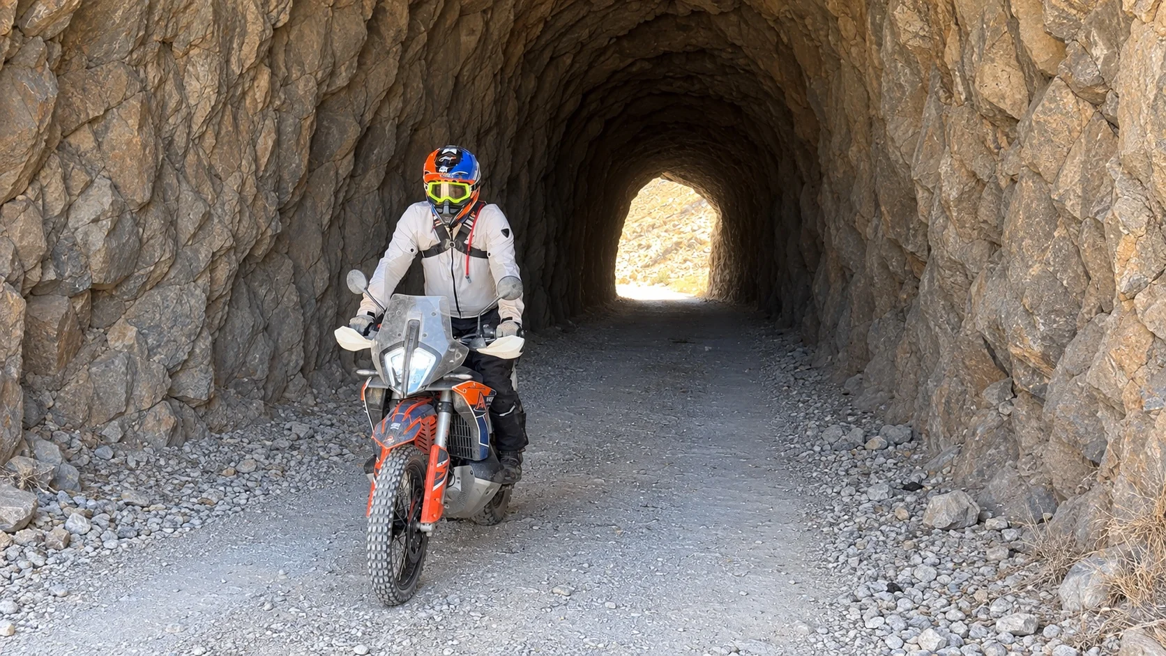 Rider on a KTM adventure bike riding out of a rock-cut tunnel on a gravel track